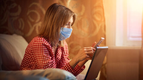 Junge Frau mit Maske und Smartphone sitzt vor einem Laptop