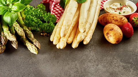 Spargel, Erdbeeren, Kartoffeln und Kräuer liegen nebeneinander auf einem dunklen Tisch