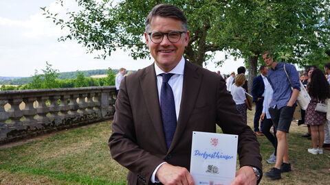 Ministerpräsident Boris Rhein mit der Urkunde „Die besten Dorfgasthäuser in Hessen“ in den Händen lächelt in die Kamera.