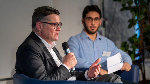 Ministerpräsident Rhein spricht in ein Mikrofon, während er in einem Sessel sitzt, hinter ihm ein Moderator.