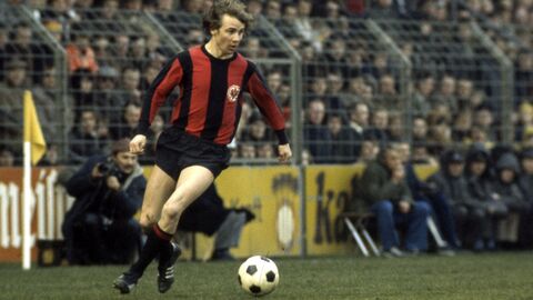Bernd Hölzenbein auf dem Fußballfeld in einem voll besetzten Stadion, neben ihm der Fußball auf dem Rasen.